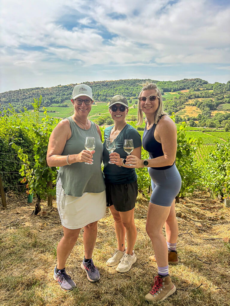 three female hiking companions in front of vineyard with glasses of wine